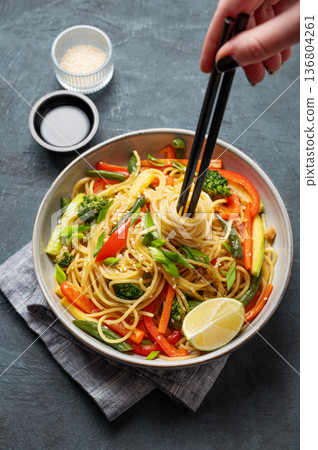 Hand holding chopsticks in plate of vegetarian wok noodles with vegetables on a dark background 136804261