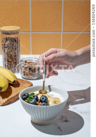 Hand holding spoon in greek yogurt with granola, blueberry and banana in a bowl on a marble table Hand holding spoon in greek yogurt with granola, blueberry and banana in a bowl on a marble table 136804269