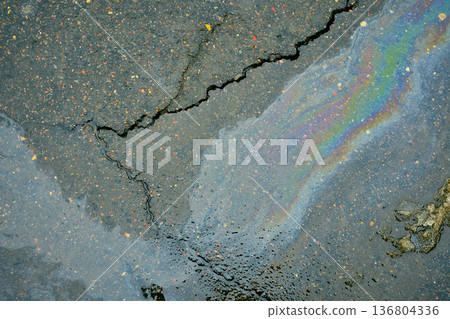 Iridescent oil slick on cracked wet asphalt surface showing rainbow colors and liquid texture. 136804336
