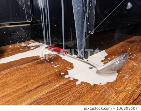 Spilled milk puddle on wooden floor with metal ladle. White liquid splashing on black kitchen cabinets. Messy accident concept 136805507