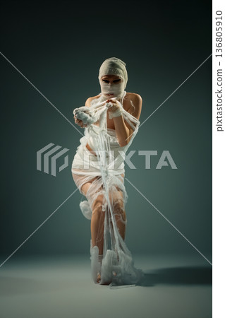 A woman wrapped in white bandages like a mummy, posing dramatically in a studio. 136805910