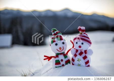 Two small funny toys baby snowman in knitted hats and scarves in deep snow outdoors on blurred mountains landscape background. Happy New Year and Merry Christmas greeting card theme. 136806287