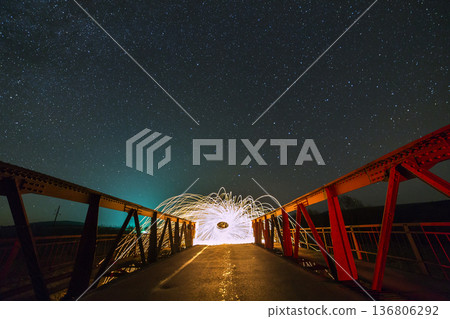 Light painting art concept. Long exposure shot of spinning steel wool in abstract circle making firework showers of bright yellow glowing sparkles on long bridge on blue night starry sky background. 136806292