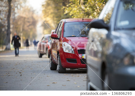 Close-up front view detail of bright red shiny car parked on pavement on background of blurred silhouette of walking people on sunny summer day. Modern city lifestyle, parking problems concept. 136806293