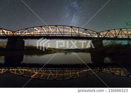 Illuminated metal bridge on concrete supports reflected in water on dark starry sky with Milky Way constellation background. Night photography concept. Illuminated metal bridge on concrete supports reflected in water on dark starry sky with Milky Way constellation background. Night photography concept. 136806323
