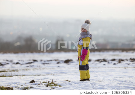 Little child girl standing outdoors alone on snow covered winter field. 136806324