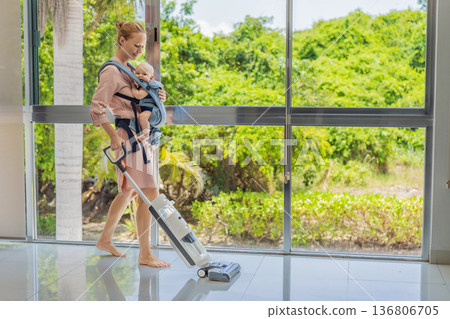 Woman vacuuming a room while carrying her baby in an ergonomic carrier. Modern motherhood, multitasking parent, home chores with infant, balance of childcare and housework concept. Woman vacuuming a room while carrying her baby in an ergonomic carrier. Modern motherhood, multitasking parent, home chores with infant, balance of childcare and housework concept. 136806705