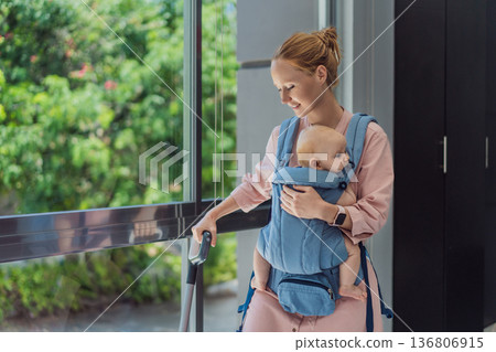 Woman vacuuming a room while carrying her baby in an ergonomic carrier. Modern motherhood, multitasking parent, home chores with infant, balance of childcare and housework concept. Woman vacuuming a room while carrying her baby in an ergonomic carrier. Modern motherhood, multitasking parent, home chores with infant, balance of childcare and housework concept. 136806915