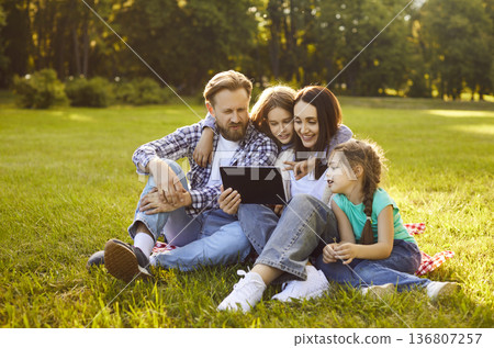 Happy family with children using tablet browsing internet sitting on grass in summer park 136807257