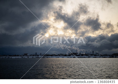 冬天的河面上,陽光透過厚厚的雲層灑下光芒:對岸是白雪皚皚的城市景觀和工業區。 冬天的河面上,陽光透過厚厚的雲層灑下光芒:對岸是白雪皚皚的城市景觀和工業區。 136808435