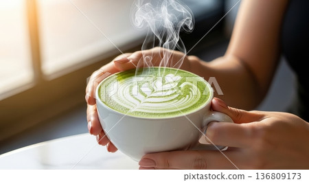A woman relaxing in a cafe with natural light pouring in, enjoying a steaming hot matcha latte with both hands. 136809173