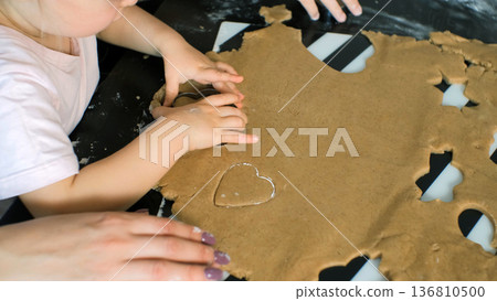 A mother gently helps her young daughter cut ginger cookie shapes from dough using small cutter A mother gently helps her young daughter cut ginger cookie shapes from dough using small cutter 136810500