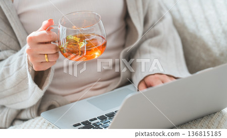 A woman sitting on a sofa drinking tea and looking at a laptop A woman sitting on a sofa drinking tea and looking at a laptop 136815185