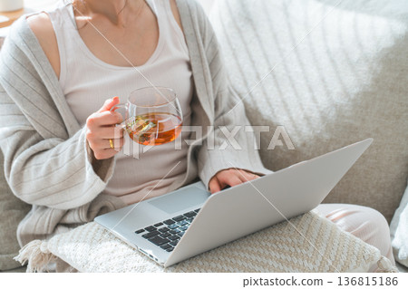 A woman sitting on a sofa drinking tea or herbal tea while looking at a computer A woman sitting on a sofa drinking tea or herbal tea while looking at a computer 136815186