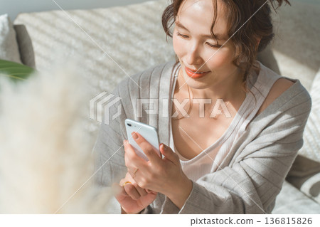 一位亞洲女性坐在客廳的沙發上，看著她的智慧型手機。 136815826