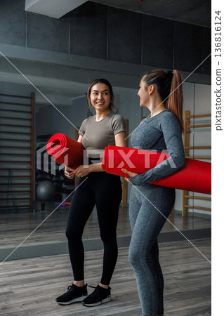 Two women hold red yoga mats in a gym while preparing for training and discussing their fitness plans 136816124