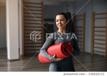 Woman holds red yoga mat in gym while preparing for fitness training session 136816131