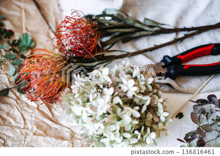 Flowers and florist tools rest on wrinkled paper and light fabric. hybrid floral design, mixed textures, dried and fresh flowers, botanical contrast, modern floristry, floral preservation Flowers and florist tools rest on wrinkled paper and light fabric. hybrid floral design, mixed textures, dried and fresh flowers, botanical contrast, modern floristry, floral preservation 136816461