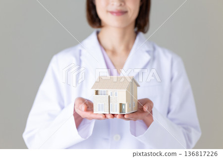 A middle-aged female doctor in a white coat holding a house model 136817226