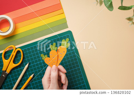 Hand holding three handmade paper carrots over green cutting mat. DIY craft project on beige background. Copyspace 136820102