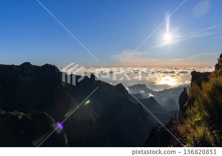 Sunburst rises above cloud sea from Pico do Arieiro with jagged peaks lens flare and shrubs edge Sunburst rises above cloud sea from Pico do Arieiro with jagged peaks lens flare and shrubs edge 136820851