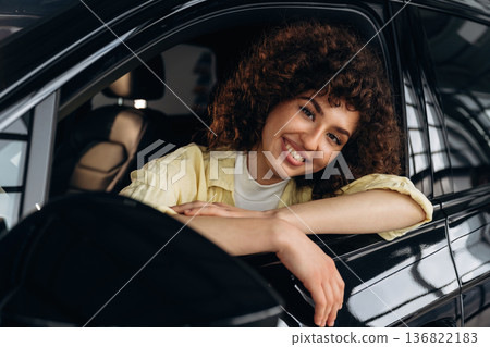 Looking through the opened window and smiling. Happy woman is sitting in the car Looking through the opened window and smiling. Happy woman is sitting in the car 136822183