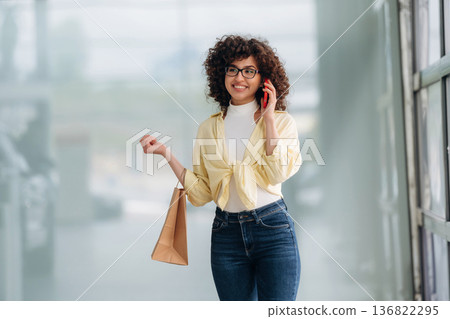 Conversation by using phone. Beautiful woman is standing indoors against blurred wall Conversation by using phone. Beautiful woman is standing indoors against blurred wall 136822295