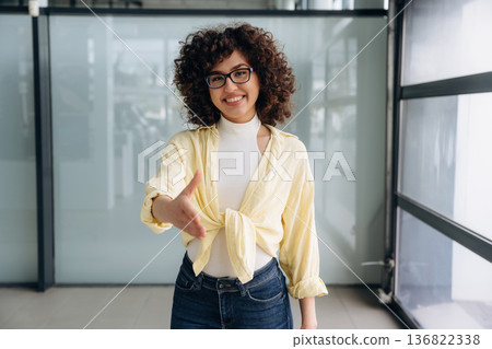Giving hand to shake. Beautiful woman is standing indoors against blurred wall 136822338