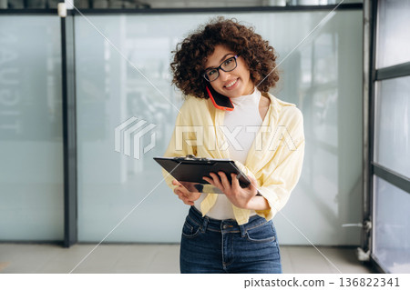 Writing the information while talking by the phone. Beautiful woman is standing indoors against blurred wall 136822341