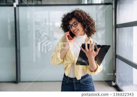 Writing the information while talking by the phone. Beautiful woman is standing indoors against blurred wall 136822343
