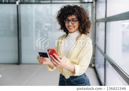 Credit card and phone in hands. Beautiful woman is standing indoors against blurred wall Credit card and phone in hands. Beautiful woman is standing indoors against blurred wall 136822346