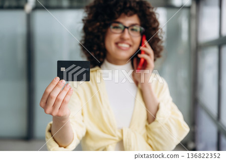 Internet payment by credit card, holding it and phone. Beautiful woman is standing indoors against blurred wall Internet payment by credit card, holding it and phone. Beautiful woman is standing indoors against blurred wall 136822352