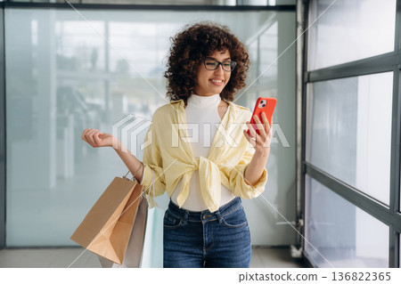 The shopping bag and phone are in hands. Beautiful woman is standing indoors against blurred wall The shopping bag and phone are in hands. Beautiful woman is standing indoors against blurred wall 136822365
