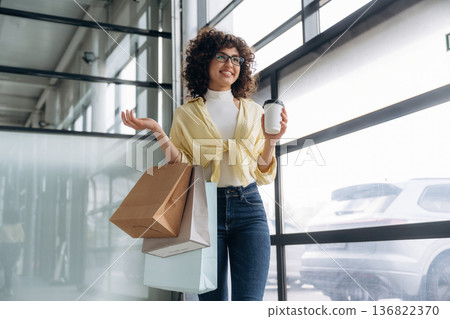 Cup of drink and shopping bags are in hands. Beautiful woman is standing indoors against blurred wall Cup of drink and shopping bags are in hands. Beautiful woman is standing indoors against blurred wall 136822370
