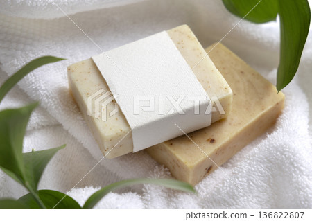 Mockup of beige solid soap bar with unbranded paper label near green leaves on white bath towels 136822807