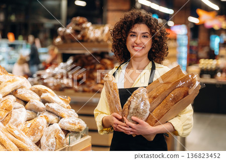 Female worker in apron is with bakery in hands, standing in the supermarket 136823452