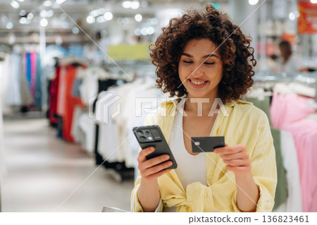 Curly hair, smile, credit card and smartphone. Woman is shopping in the clothing store Curly hair, smile, credit card and smartphone. Woman is shopping in the clothing store 136823461