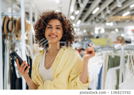 Curly hair, smile, credit card and smartphone. Woman is shopping in the clothing store 136823592
