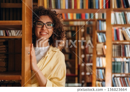 Hides and peeks from behind the shelf. Beautiful woman is in the library 136823664