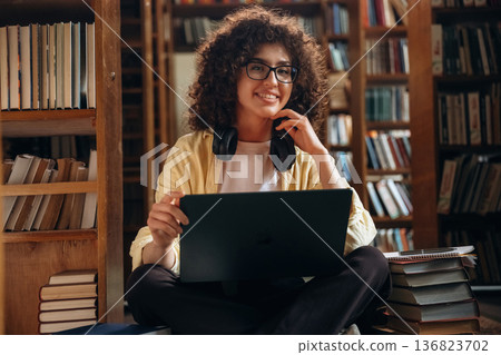 In headphones and with laptop, sitting. Beautiful woman is in the library In headphones and with laptop, sitting. Beautiful woman is in the library 136823702