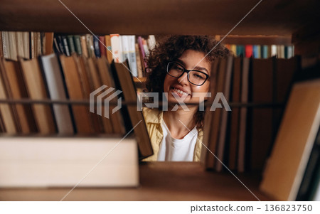 Searching for the right book, view through the shelf. Beautiful woman is in the library Searching for the right book, view through the shelf. Beautiful woman is in the library 136823750