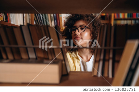 Searching for the right book, view through the shelf. Beautiful woman is in the library Searching for the right book, view through the shelf. Beautiful woman is in the library 136823751