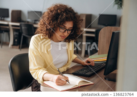 Making information research, with notepad and pen. Woman is in the library section with computers Making information research, with notepad and pen. Woman is in the library section with computers 136823764