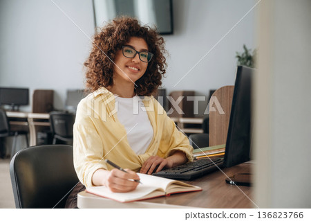 By the table with pen and notepad. Woman is in the library section with computers By the table with pen and notepad. Woman is in the library section with computers 136823766