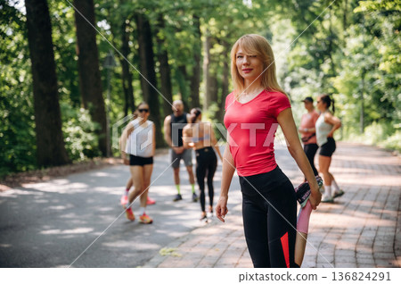 Blonde woman is standing in front of people. Group of runners are together outdoors Blonde woman is standing in front of people. Group of runners are together outdoors 136824291
