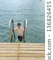 Smiling teenage boy climbing a ladder out of the sea onto a wooden pier. 136826455