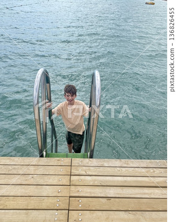 Smiling teenage boy climbing a ladder out of the sea onto a wooden pier. Smiling teenage boy climbing a ladder out of the sea onto a wooden pier. 136826455