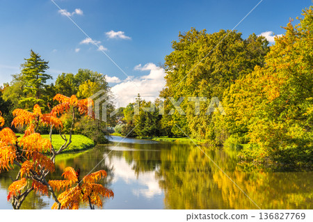 Idyllic autumn scene features a tranquil river mirroring colorful trees under a clear blue sky with scattered clouds. Lednice Castle garden in South Moravia region of the Czech Republic, Europe. Idyllic autumn scene features a tranquil river mirroring colorful trees under a clear blue sky with scattered clouds. Lednice Castle garden in South Moravia region of the Czech Republic, Europe. 136827769