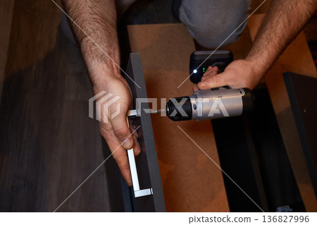 Person uses a power drill to assemble furniture in a room during daylight hours in a home setting Person uses a power drill to assemble furniture in a room during daylight hours in a home setting 136827996