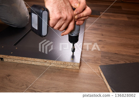 A person is installing a part of furniture by using a power drill. The setting shows wooden floors. A person is installing a part of furniture by using a power drill. The setting shows wooden floors. 136828002